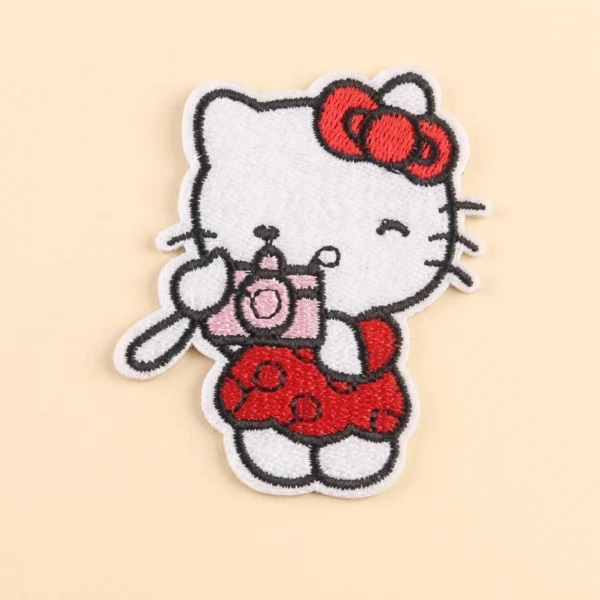 Cartoon cat embroidered iron-on patch with a red bow and pink camera on a cream background.